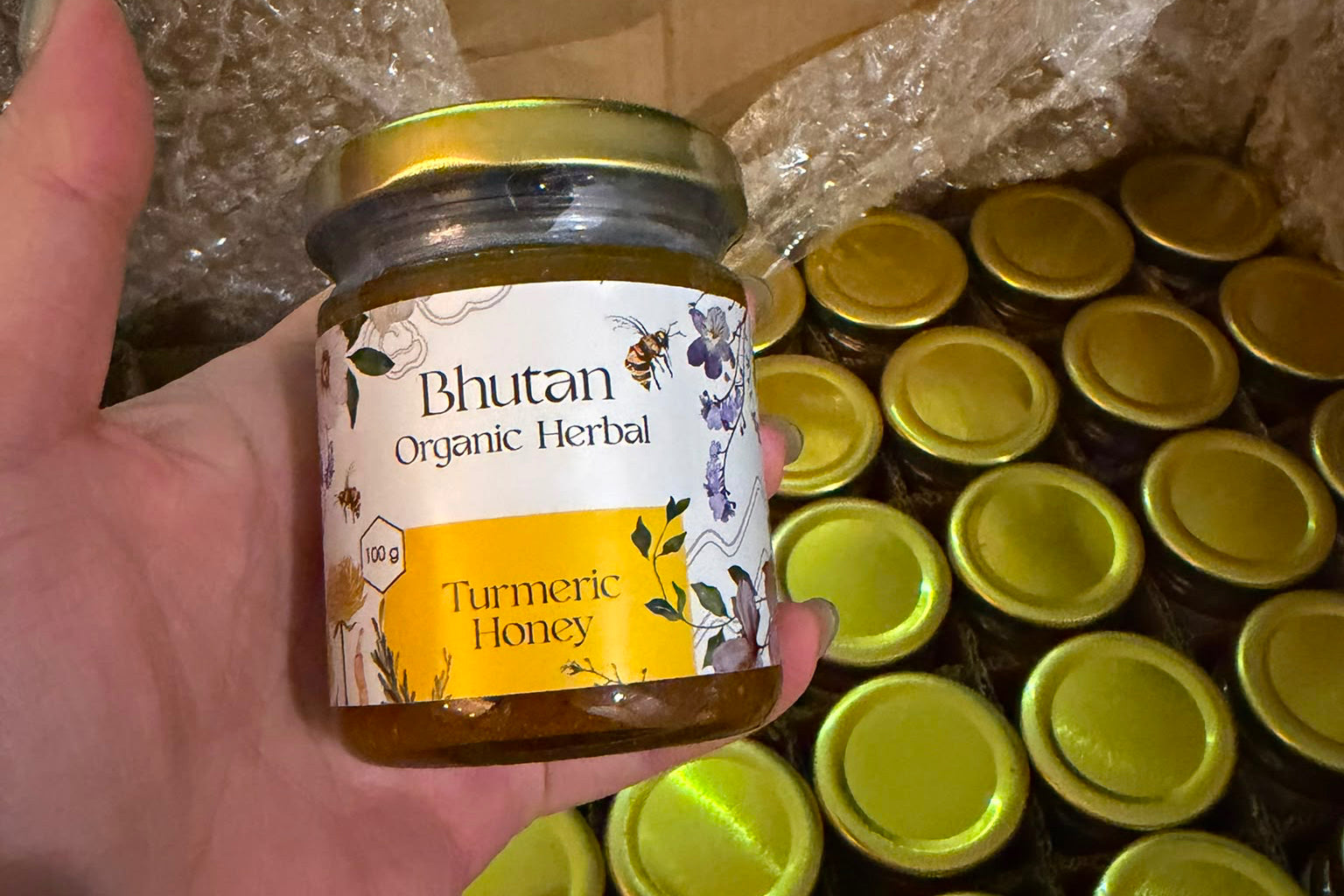 A hand holds a glass jar of Bhutan Organic Herbal Turmeric Honey, with a cardboard box filled with multiple identical jars in bubble wrap in the background.
