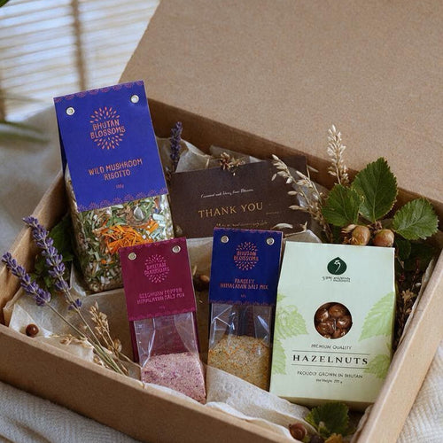 A curated gift box filled with packaged food items, including a box of hazelnuts, a purple tea packet, a pink tea packet, and a dark thank-you note, arranged in a brown cardboard box on a white cloth over a wooden surface.