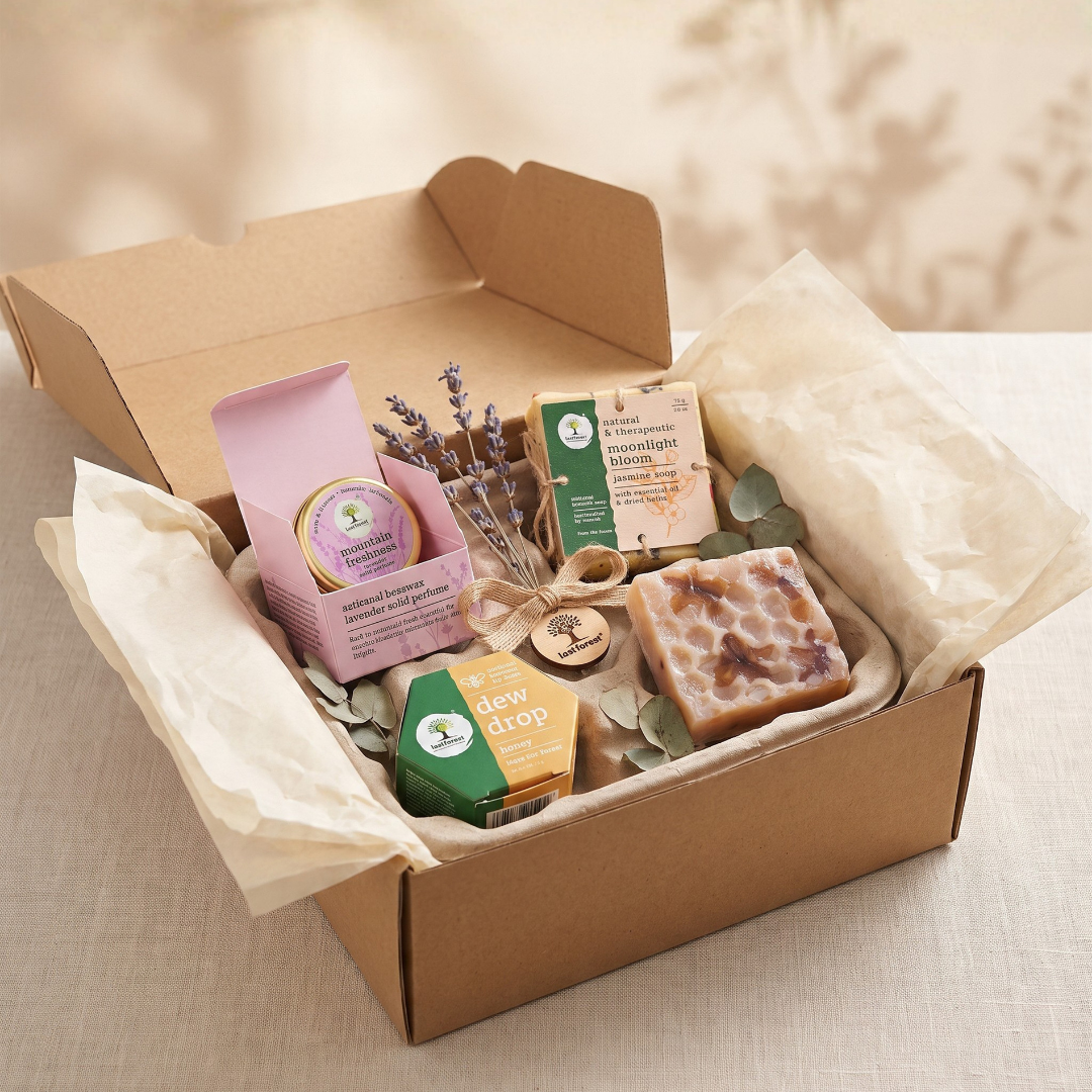 A curated gift box filled with artisanal soap bars, a pink packaged item, a small green soap, dried lavender sprigs, and eucalyptus leaves, arranged in a kraft cardboard box lined with cream tissue paper on a neutral fabric surface.