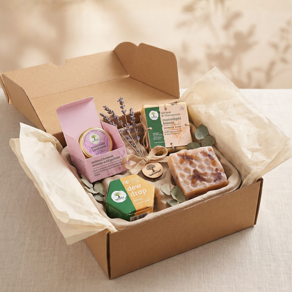 A curated gift box filled with artisanal soap bars, a pink packaged item, a small green soap, dried lavender sprigs, and eucalyptus leaves, arranged in a kraft cardboard box lined with cream tissue paper on a neutral fabric surface.