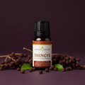 A bottle of Kingdom Essentials Thingye essential oil surrounded by dried berries and green leaves on a dark purple background.