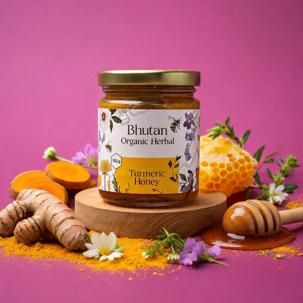A jar of Bhutan Organic Herbal Turmeric Honey sits on a round wooden pedestal, surrounded by fresh turmeric roots, sliced turmeric, a honeycomb, a honey dipper, and small white and purple flowers against a solid pink background.